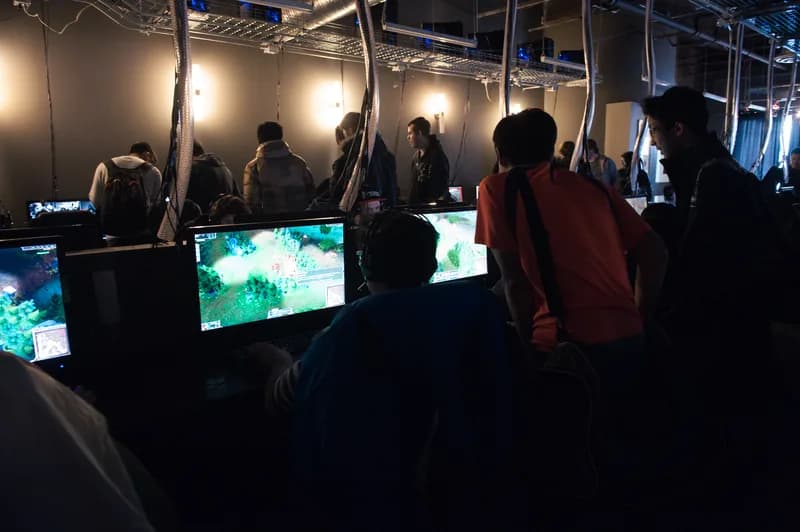 Moody row of glowing game screens with silhouetted players and spectators at a LoL tournament